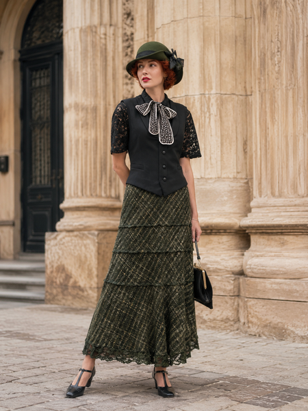 „Elegantes 20er Jahre Damen Outfit mit schwarzer Weste, Spitzenbluse, gemustertem Stufenrock und grünem Vintage Hut vor historischer Architektur“