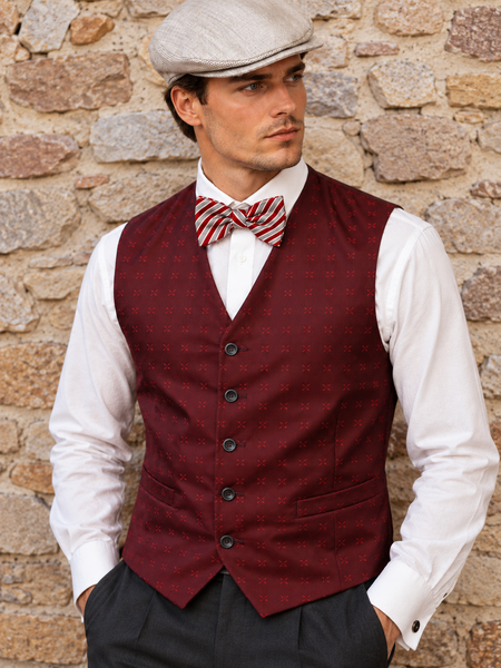 20er Jahre Herren Outfit mit bordeauxroter Weste, gestreifter Fliege und heller Schiebermütze vor Steinwand