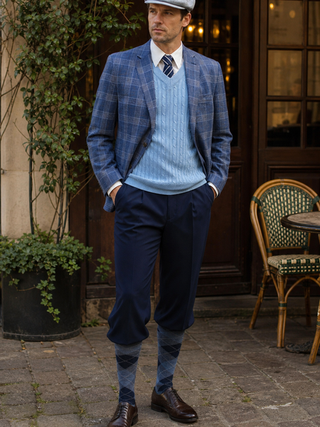 Herren im hochwertigen 20er Jahre Outfit mit blau kariertem Sakko, hellblauem Zopfmuster-Pullover, Knickerbockerhose, Rautenstrümpfen und Schiebermütze vor einem Café.