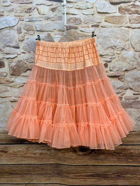 „Apricotfarbener Petticoat an Kleiderbügel hängend vor Steinwand, mit verstellbarem Satinbund und mehrlagigen Tüllvolants.