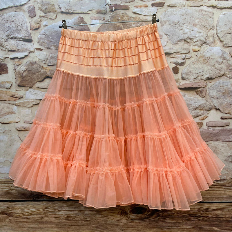 „Apricotfarbener Petticoat an Kleiderbügel hängend vor Steinwand, mit verstellbarem Satinbund und mehrlagigen Tüllvolants.