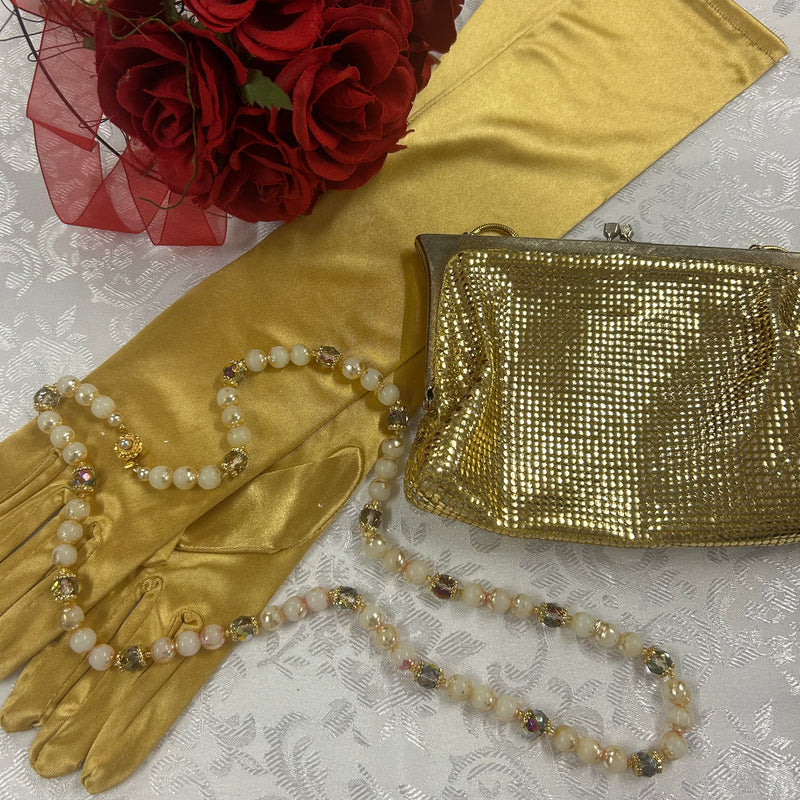 Goldfarbene Vintage-Handtasche mit Perlenkette und langen Satin-Handschuhen auf weißem Hintergrund.