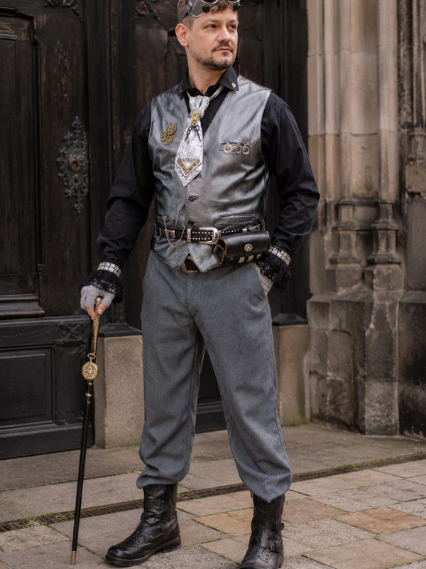 Steampunk Herren Outfit mit silberfarbener Lederweste, Goggles, Schurwollhose und Accessoires an echtem Modell vor historischem Gebäude.