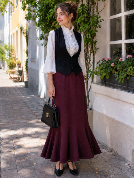 Model trägt ein Vintage Damen Outfit mit weißer Spitzenbluse, schwarzer Weste, langem bordeauxfarbenem Rock und Handtasche in einer hellen Altstadtstraße