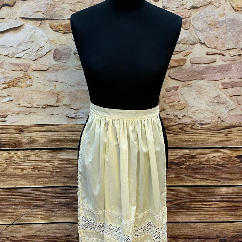 „Cremefarbene Vintage-Dirndlschürze mit Spitzenborten und Falten am Bund.“